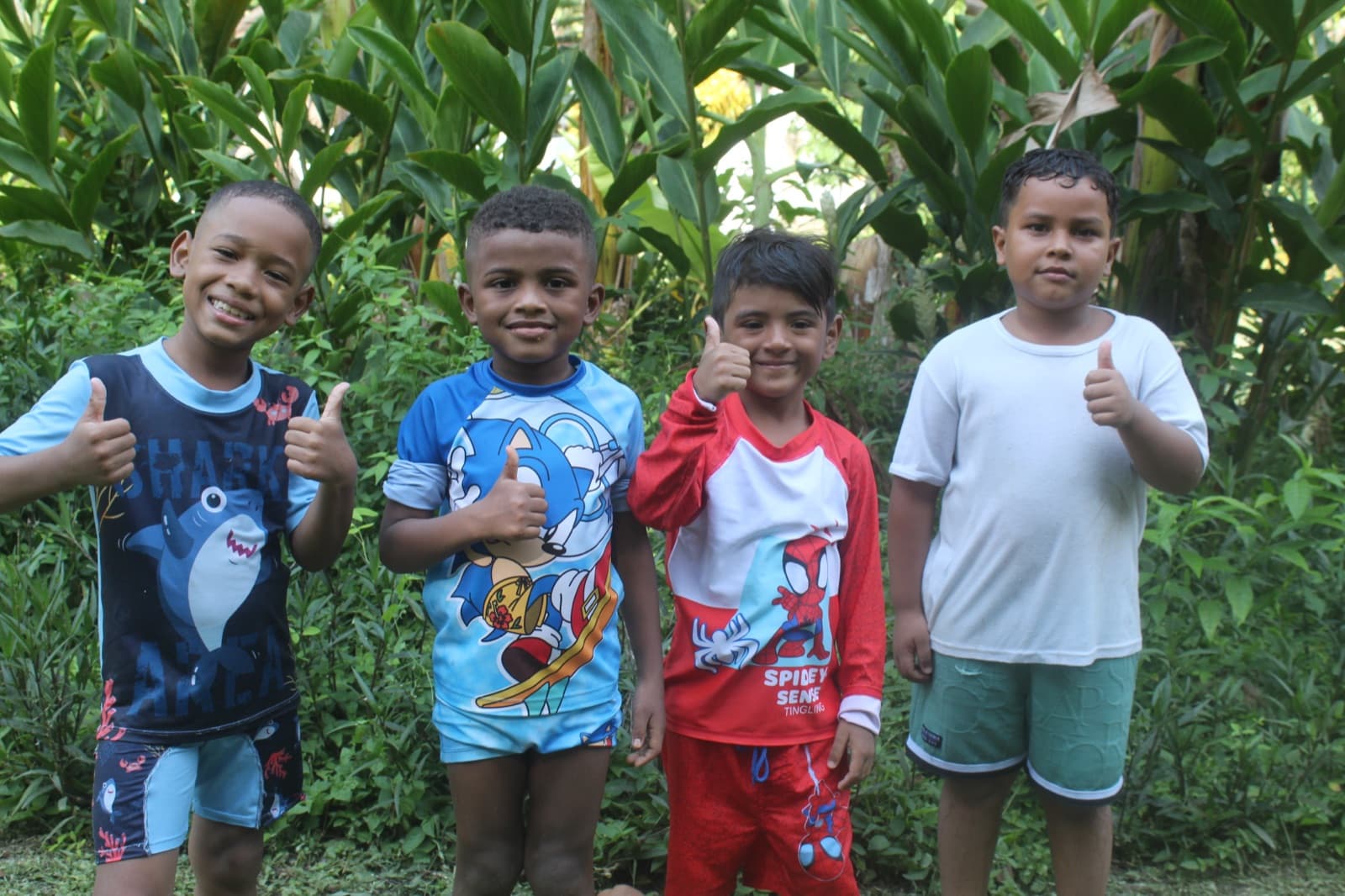 Four boys giving a thumbs up outdoors in front of green plants
